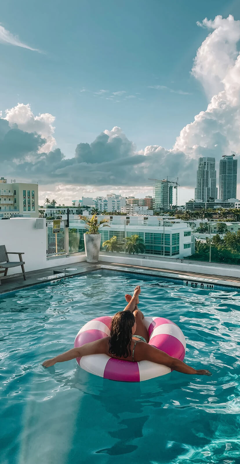 A woman relaxes on a pink and white pool float in a rooftop pool overlooking the city.