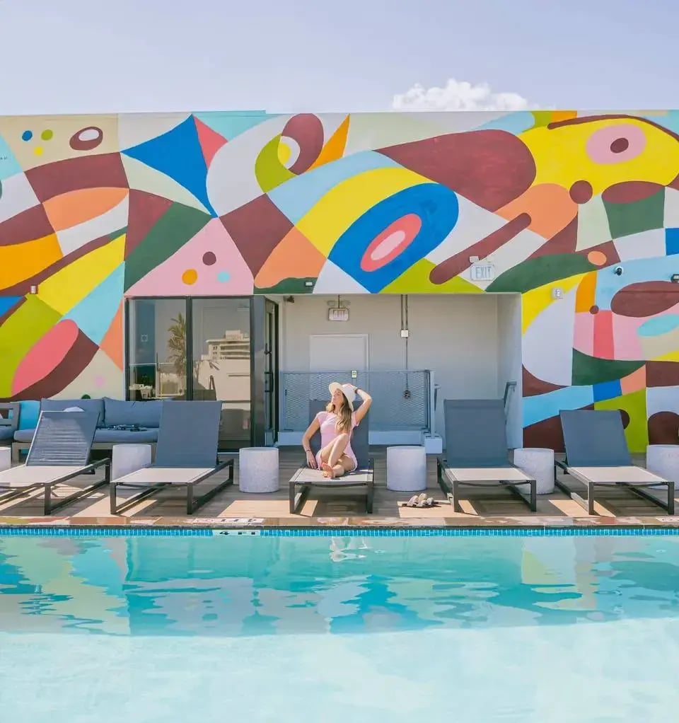 A woman sits on a lounge chair in front of a pool with a colorful mural in the background.