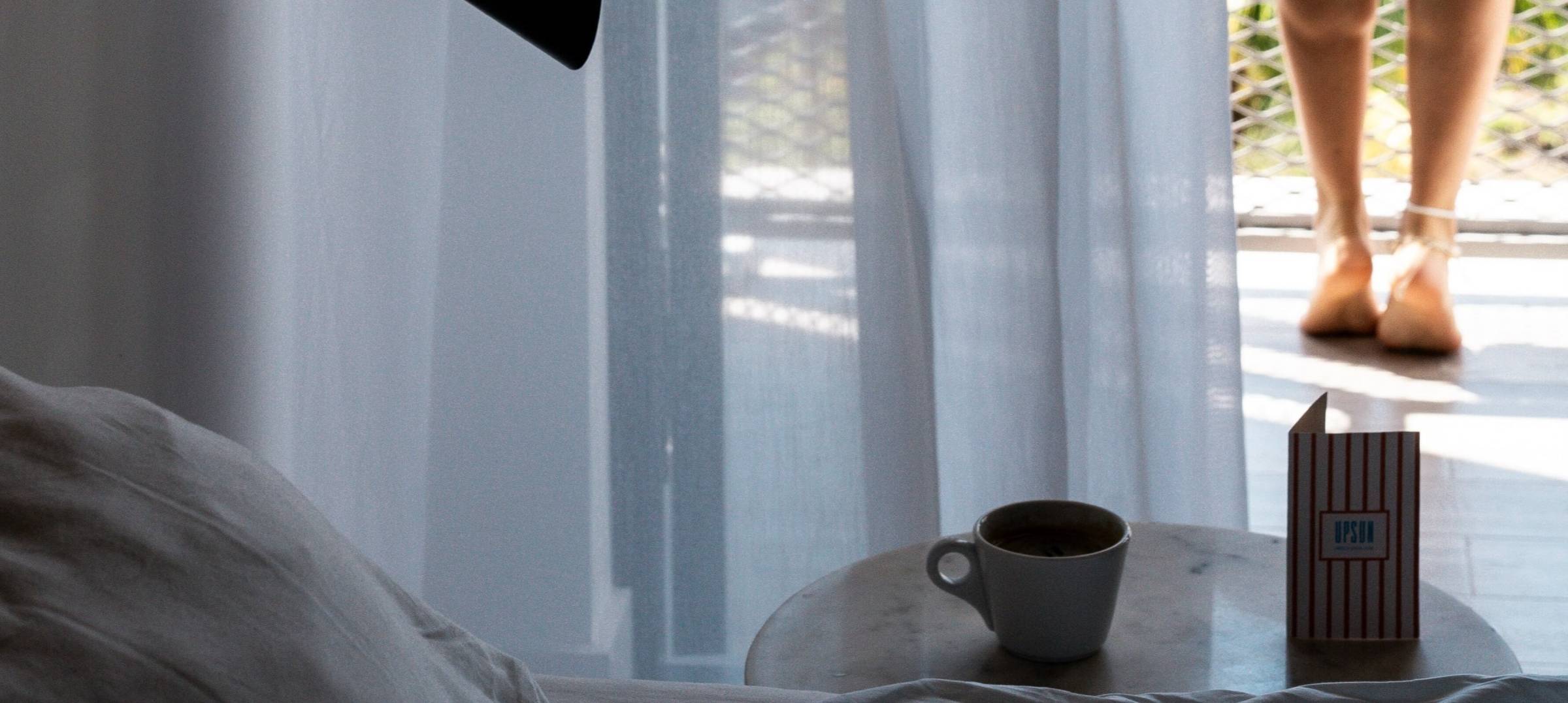 A room with a bed, a small table with a coffee cup, and a balcony with a person's legs visible outside.