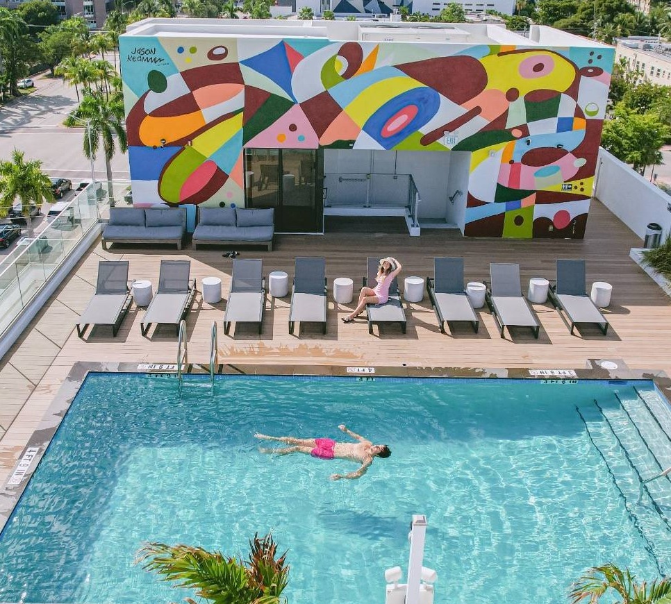Aerial view of a rooftop pool with a colorful mural on the wall, people lounging on chairs, and a man swimming.